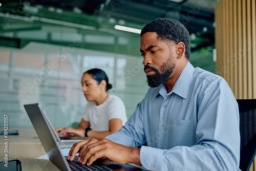 Serious male employee working diligently at laptop in a professional open-plan office setting, focusing on work with colleague