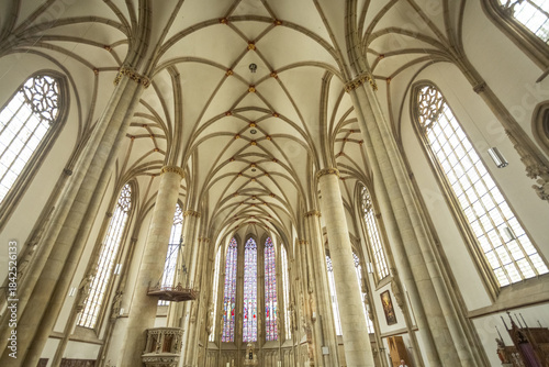 View of rows of wooden pews leading to a stained-glass window in Lambertikirche church with high vaulted ceilings and stone columns, Muenster, North Rhine-Westphalia, Germany.