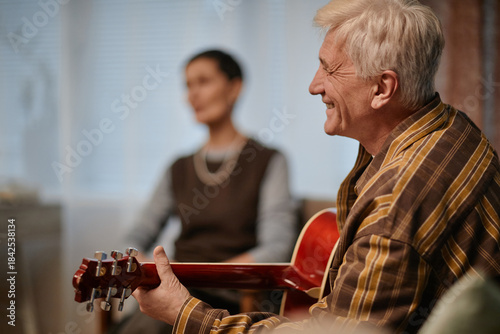 Senior Caucasian man playing acoustic guitar while smiling, sitting indoors with blurred middle aged woman in background, nursing home setting suggesting recreational group activity