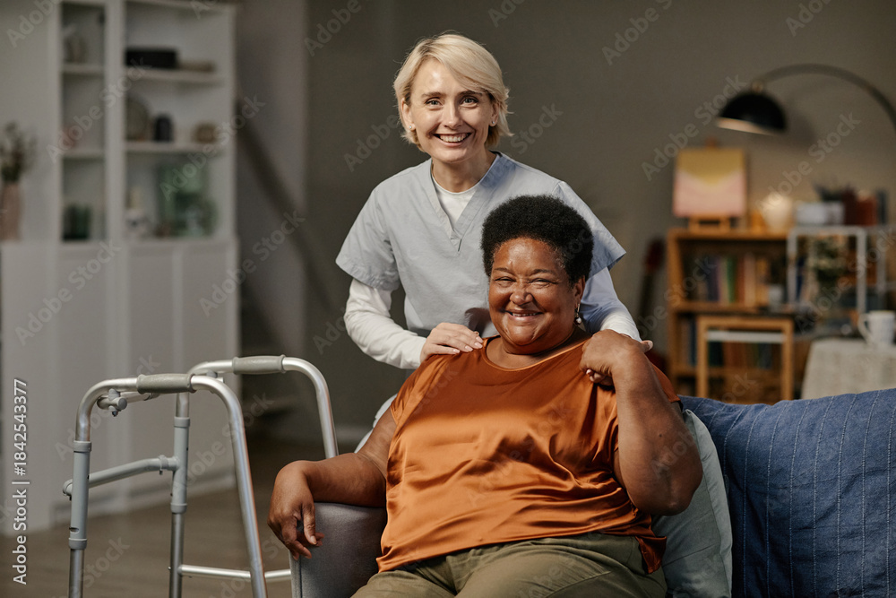 Obraz premium Caucasian middle aged woman caregiver standing behind seated Black senior woman smiling in nursing home setting, both women looking at camera, walker visible beside chair