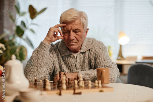Senior Caucasian man sitting at table concentrating on chess game in nursing home, resting head on hand, coffee mug nearby, thoughtful expression, natural indoor setting