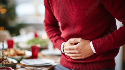 Man in a red sweater experiencing stomach discomfort after holiday meal with festive table filled with food in the background.