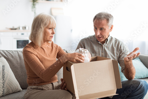 Konstfotografi A couple sits on a couch in a bright living room, opening a box together