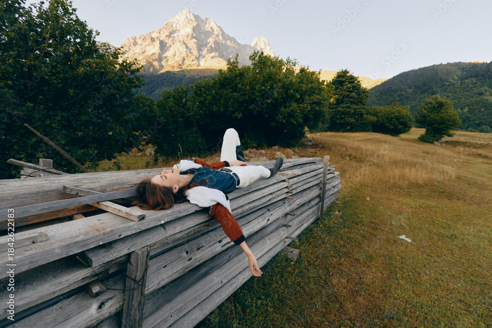 Fototapeta premium Woman lying to relax on a wooden fence in a grassy meadow beneath a mountain, nature landscape with sunlight and trees. Young female in denim vest and boots enjoying peaceful rural rest.