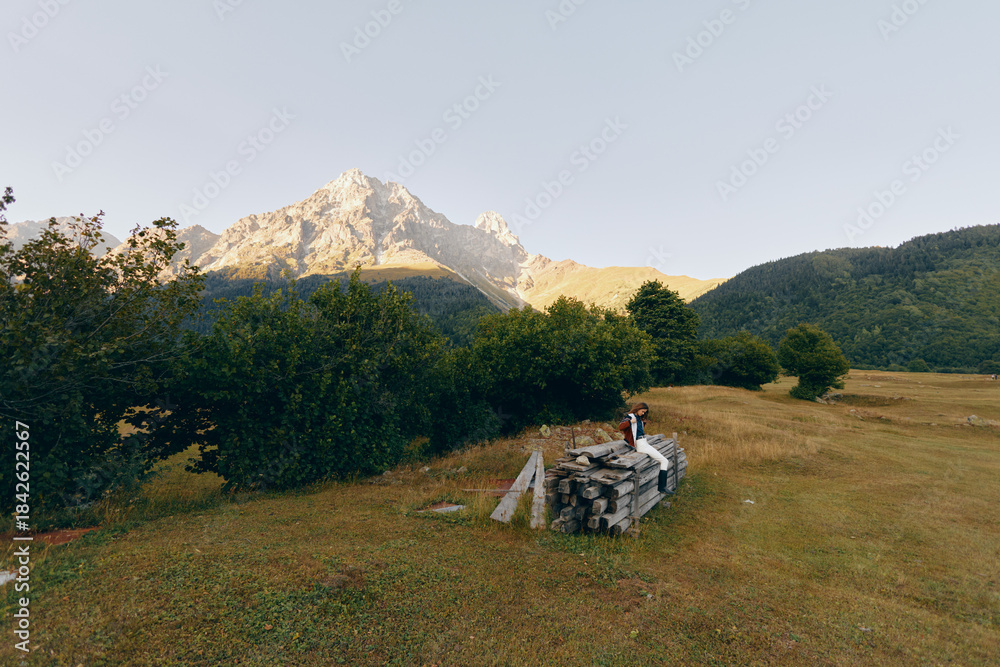 Fototapeta premium Mountain meadow with woodpile and person sitting near trees, landscape view of nature and hiking trail at sunrise, peaceful alpine field and rustic outdoor scenery.