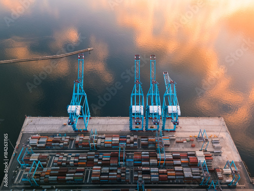 Aerial view of towering blue cranes oversee a grid of colorful shipping containers under a sky mirroring the water's surface, Rijeka, Primorje-Gorski Kotar County, Croatia.