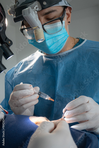 Vertical shot of a concentrated male plastic surgeon wearing loupes and a headlamp, administering an anesthesia injection to a patient during a blepharoplasty procedure in an operating room.