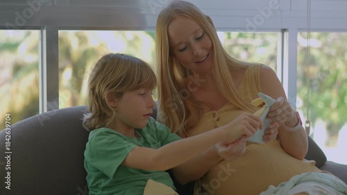 Pregnant mother and older son looking at baby clothes on the couch. Family bonding, sibling anticipation, and preparing for new baby concept for parenting blogs, maternity content and lifestyle ads