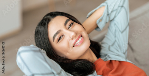 A young woman is seen lounging comfortably on a couch with her hands resting behind her head. She has a bright smile, and the warm setting creates a relaxed atmosphere.