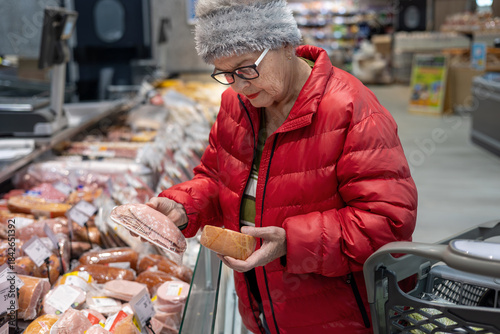 Photos Senior woman reading product label while shopping for deli meat in supermarket refrigerated section