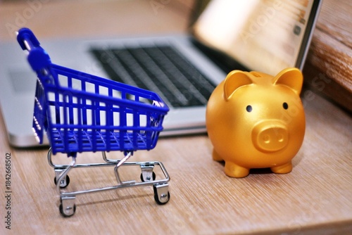 Golden piggy bank near a blue shopping cart and laptop as background. Concept of online shopping.