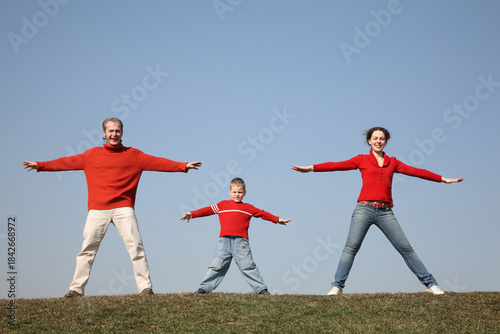 A family of three is standing on a grassy hill, with the man and boy on one side and the woman on the other. They are all wearing red shirts and are stretching their arms out to the side