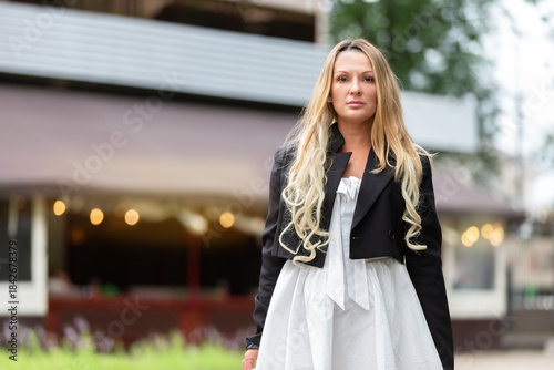Woman in white dress and black jacket stands confidently outside a cafe during daytime