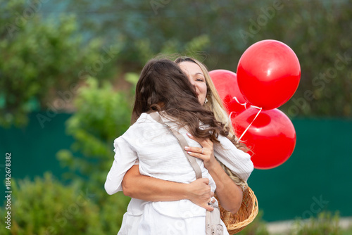 Emotional embrace between a mother and daughter with red balloons during a sunny outdoor event in spring
