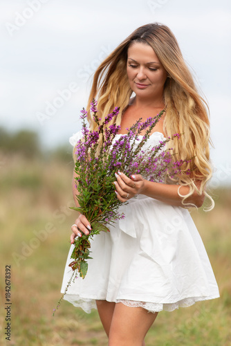 Woman in a white dress walking through a field while holding a bouquet of purple wildflowers on a cloudy day