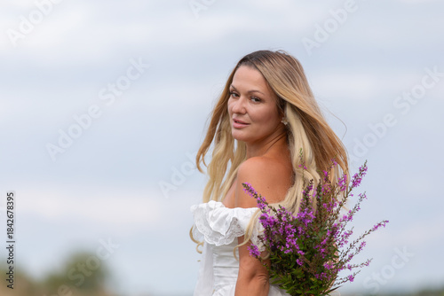 Woman in a white dress holds purple flowers in a field on a cloudy day, showcasing natural beauty and tranquility