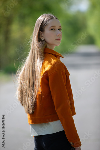 Model posing in an orange jacket while walking down a serene path surrounded by greenery during a sunny day
