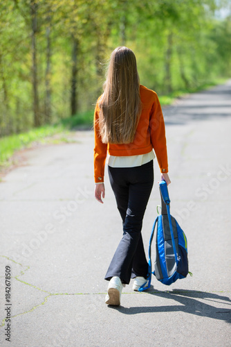 Young woman walks down a quiet path in nature during a sunny afternoon, carrying a blue backpack and wearing a stylish orange jacket