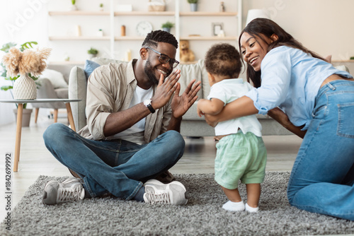 Billede på lærred In a warm living room, a baby boy joyfully takes his first steps while his parents cheer him on