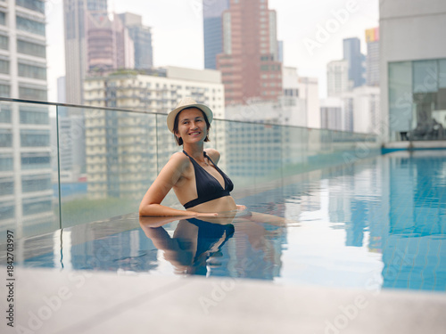 Photography Young asian woman relaxing by pool at Kuala Lumpur hotel with view of surrounding skyscrapers, enjoying leisure time in vibrant urban setting
