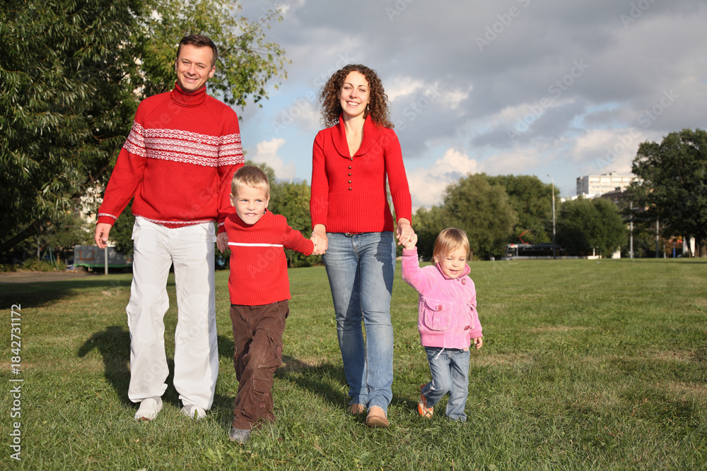 Obraz premium A family of four is walking in a park, with a man and a woman holding hands and a child in each of their arms. The family is wearing red clothing, and the atmosphere is warm and joyful