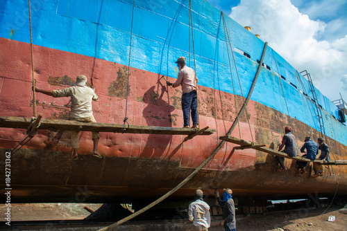 Dhaka, Bangladesh - 31 August 2024: View of workers precariously perched on scaffolding, painting a colossal ship's hull with vibrant blue and red hues.