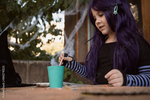 Little girl wearing purple wig and witch costume smiling happily and painting by paint and brush