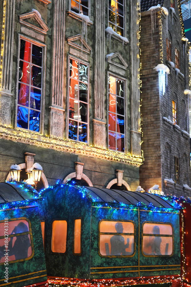 Obraz premium Colorful festive train decorated with bright lights is parked in front of a beautifully illuminated building, creating a magical winter wonderland atmosphere for holiday celebrations