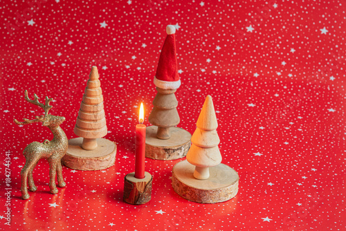 burning red candle with wooden trees around on wooden coasters, Santa hat and golden dear on red background with white stars