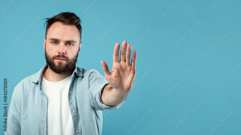 Naklejka premium A man with a beard and short hair stands against a solid blue background. He raises one hand, signaling for attention or stopping. His expression appears serious yet calm, creating a moment of pause.