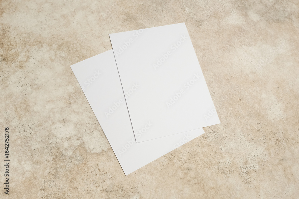 Fototapeta premium Two blank white sheets of paper stacked on a textured beige surface, ideal for design mockups, presentations, or creative projects with ample copy space for text