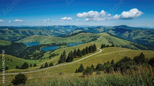 Rolling green hills and lakes under a blue sky with clouds, scenery with winding road and trees.