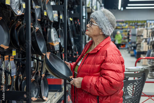 Tableau sur toile elderly woman shopping independently in a supermarket housewares section, carefully choosing a frying pan from a wide selection of cookware