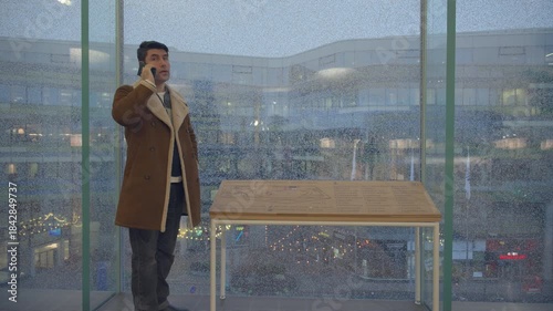 Wallpaper Mural A man in a brown shearling coat stands indoors by rain-covered windows, talking on his phone. Behind him, a softly lit shopping mall or city square is visible, with a wooden information table nearby Torontodigital.ca