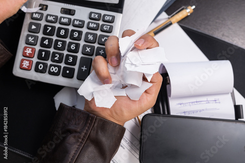 Fotografia Man sitting at an office desk destroying bills and financial documents, expressing intense stress and anger caused by debt, surrounded by work tools and paperwork