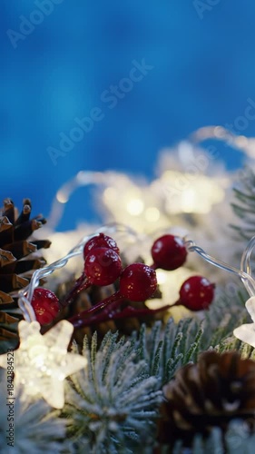 An extreme close-up video of a flickering candle flame, golden light on frosted fir branches, sparkling snow, and pine cones. A smooth push-in reveals red berries, star lights, and a shimmering blue