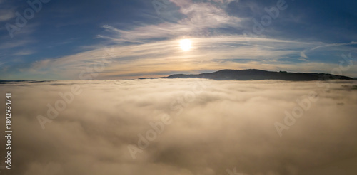 Sun above a sea of fog and clouds with mountain ridge under blue sky, aerial view of foggy mountains