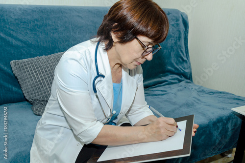 Female doctor in white coat with stethoscope sitting on sofa and writing notes on clipboard after home visit, documenting patient symptoms, treatment plan and follow up recommendations