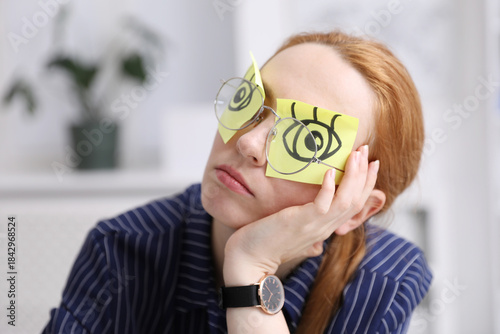 Woman with fake eyes drawn on sticky notes snoozing at workplace in office, closeup