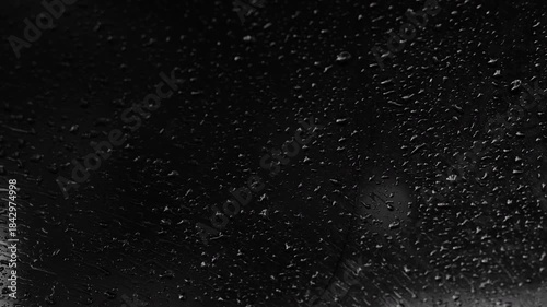 Extreme close-up cinematic black and white view of raindrops on a car window at night. Moody, abstract and atmospheric textural footage with high contrast detail
