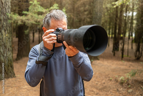 Older man enjoying birdwatching hobby, using camera with telephoto lens in forest