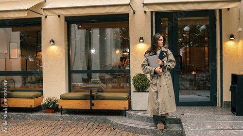 Cuadro en lienzo Young woman in trench coat holding laptop walks down steps outside a modern cafe