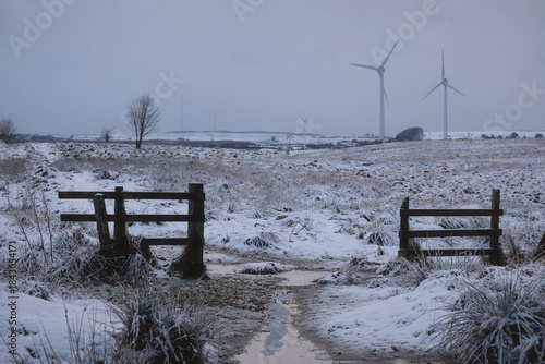 Winter rural landscape with a snow covered path, wooden gates and wind turbines in the distance. Cold countryside scene showing renewable energy and seasonal weather.