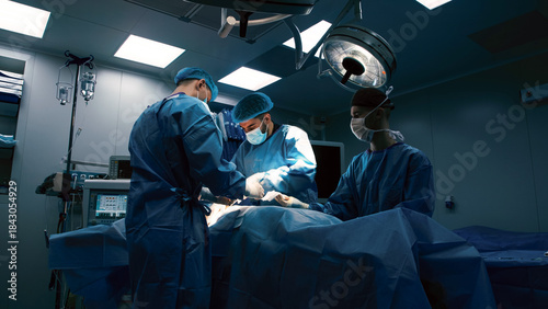 Surgical team of three medical professionals in blue scrubs and masks performing an operation in a well-lit operating room with advanced medical equipment and sterile environment