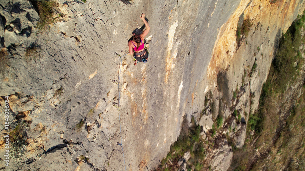 Obraz premium AERIAL: View above a brave female climber high on an exposed limestone face. Young woman lead climbing a demanding route in picturesque Cikola canyon. Adrenaline climbs in Croatian climbing area.