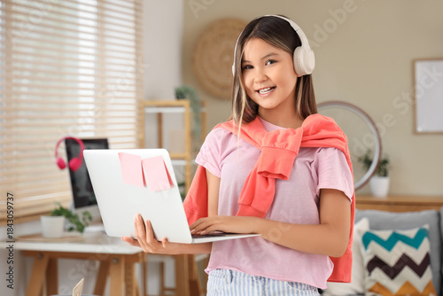 Smiling school girl wearing wireless headphones and using laptop at home, hap...