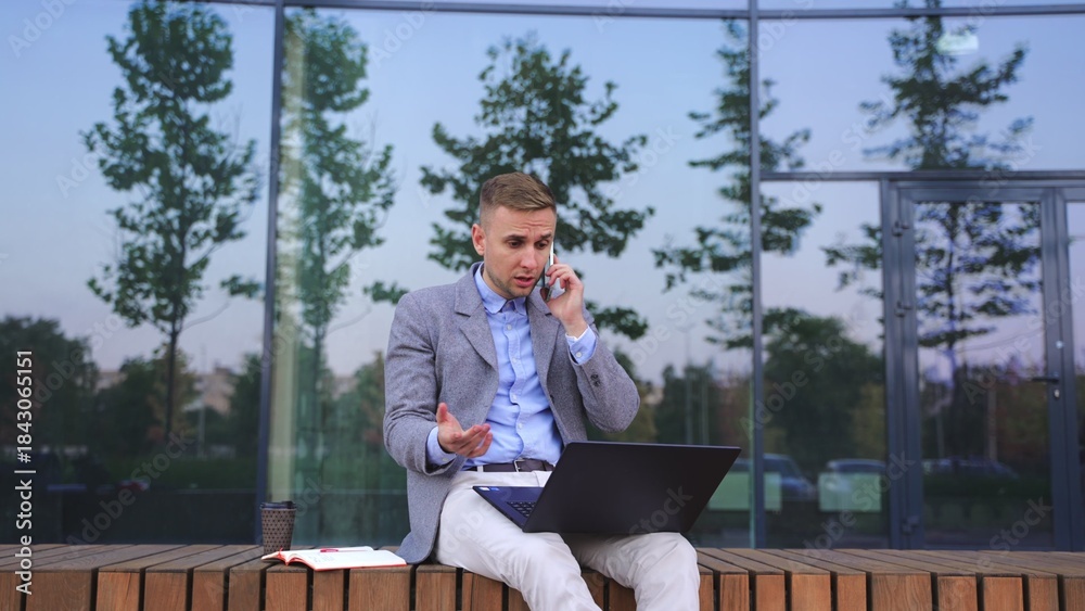 Fototapeta premium Angry man working with laptop, talking on the phone, sit on bench. Slow motion
