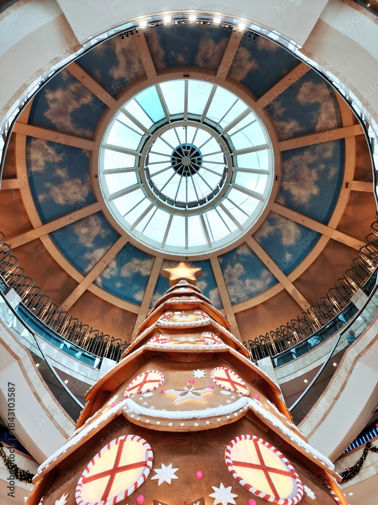 Obraz premium A striking, vertical, low-angle shot of a giant gingerbread-themed Christmas tree dominating the atrium of a grand shopping mall, crowned by a large, circular skylight with a painted ceiling of clouds
