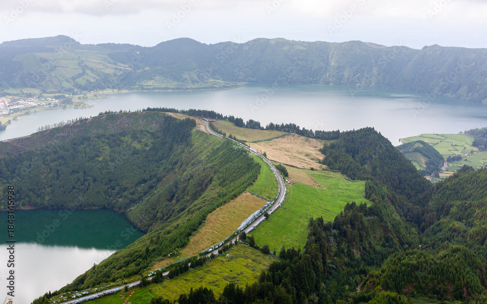 Fototapeta premium Volcanic lake in Sete Cidades, Sao Miguel, Azores. Winding road through green countryside. Caldera landscape with mountains and clouds.