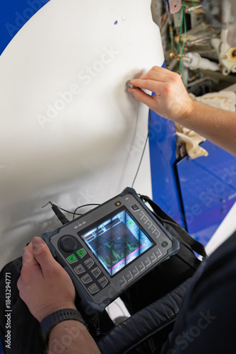 Professional NDT inspector conducting ultrasonic testing on aeroplane rudder surface with a handheld flaw detection device, showing waveform analysis for aircraft maintenance and structural integrity